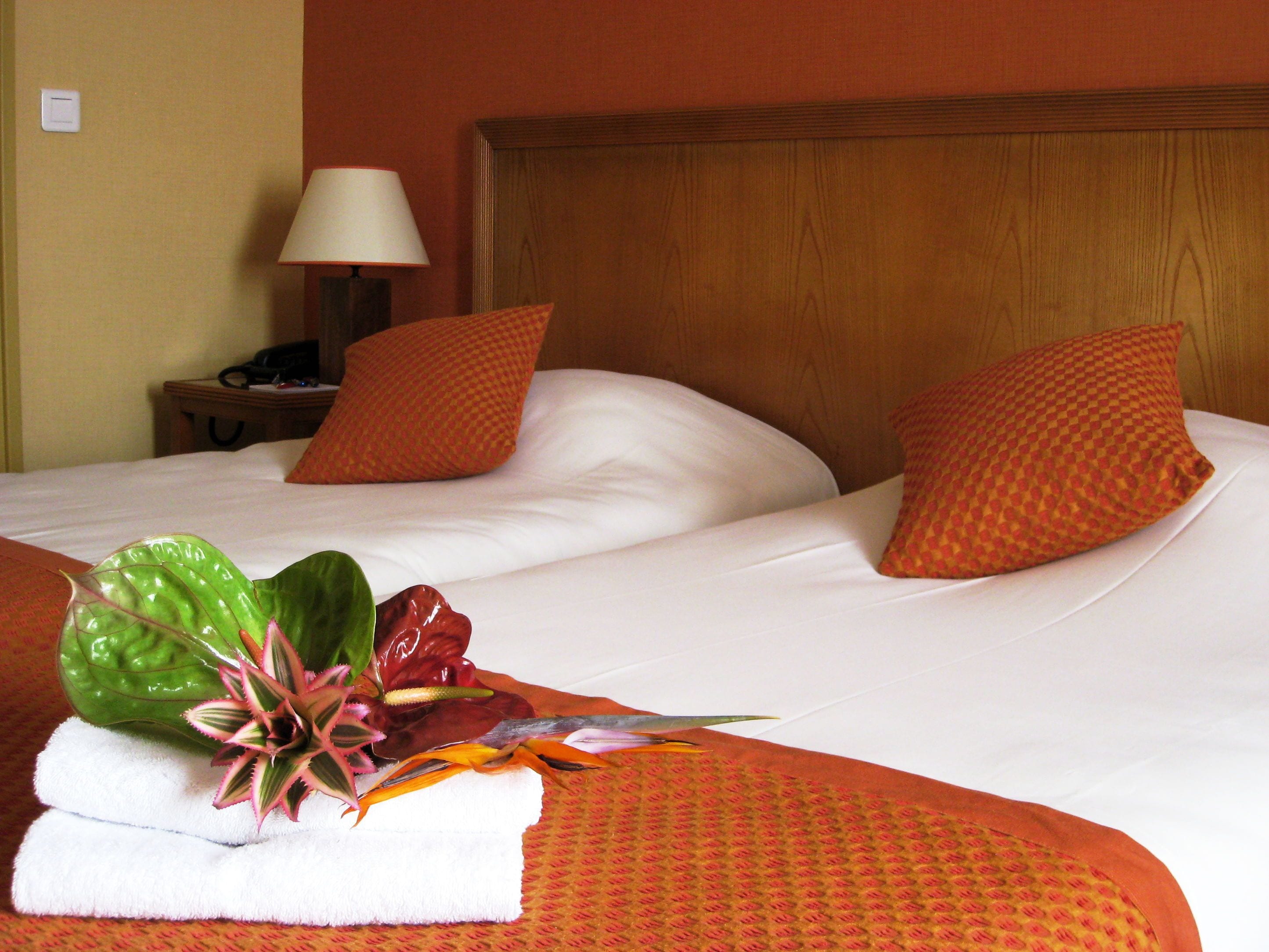 Habitación de hotel con dos camas, cada una con ropa blanca y almohadas naranjas. Cuenta con un cabecero de madera, una mesita de noche con una lámpara y un teléfono. Un arreglo decorativo de hojas y flores se coloca sobre las camas.