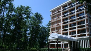 Um hotel com vários andares e entrada coberta, rodeado por vegetação exuberante e árvores.
