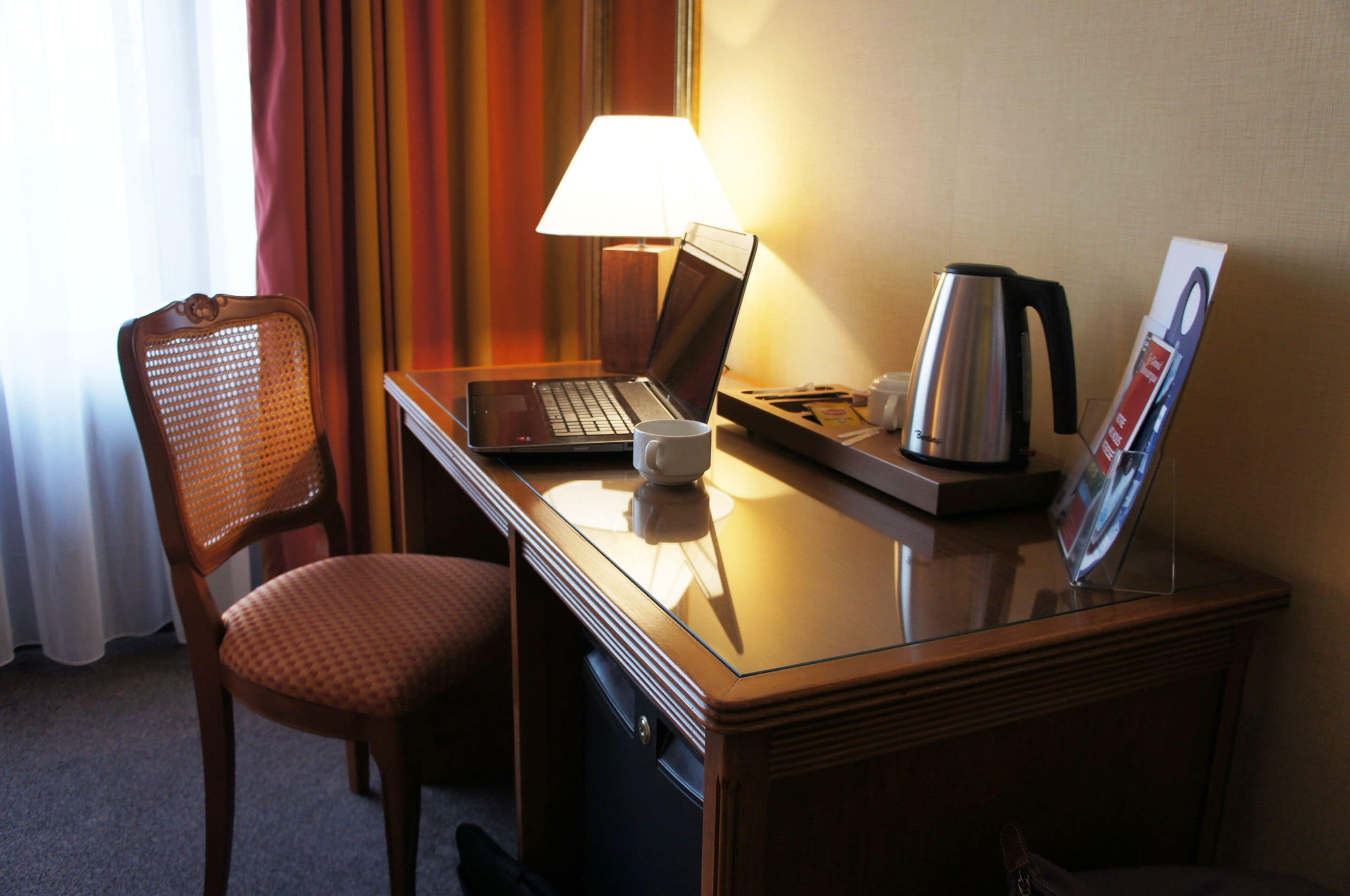 Ein Hotelzimmer mit einem Holztisch, einem Laptop, einer Kaffeemaschine, einem Sessel und einer Lampe.