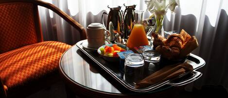 Bandeja de desayuno con frutas, jugo, café y flores sobre una mesa junto a una ventana.