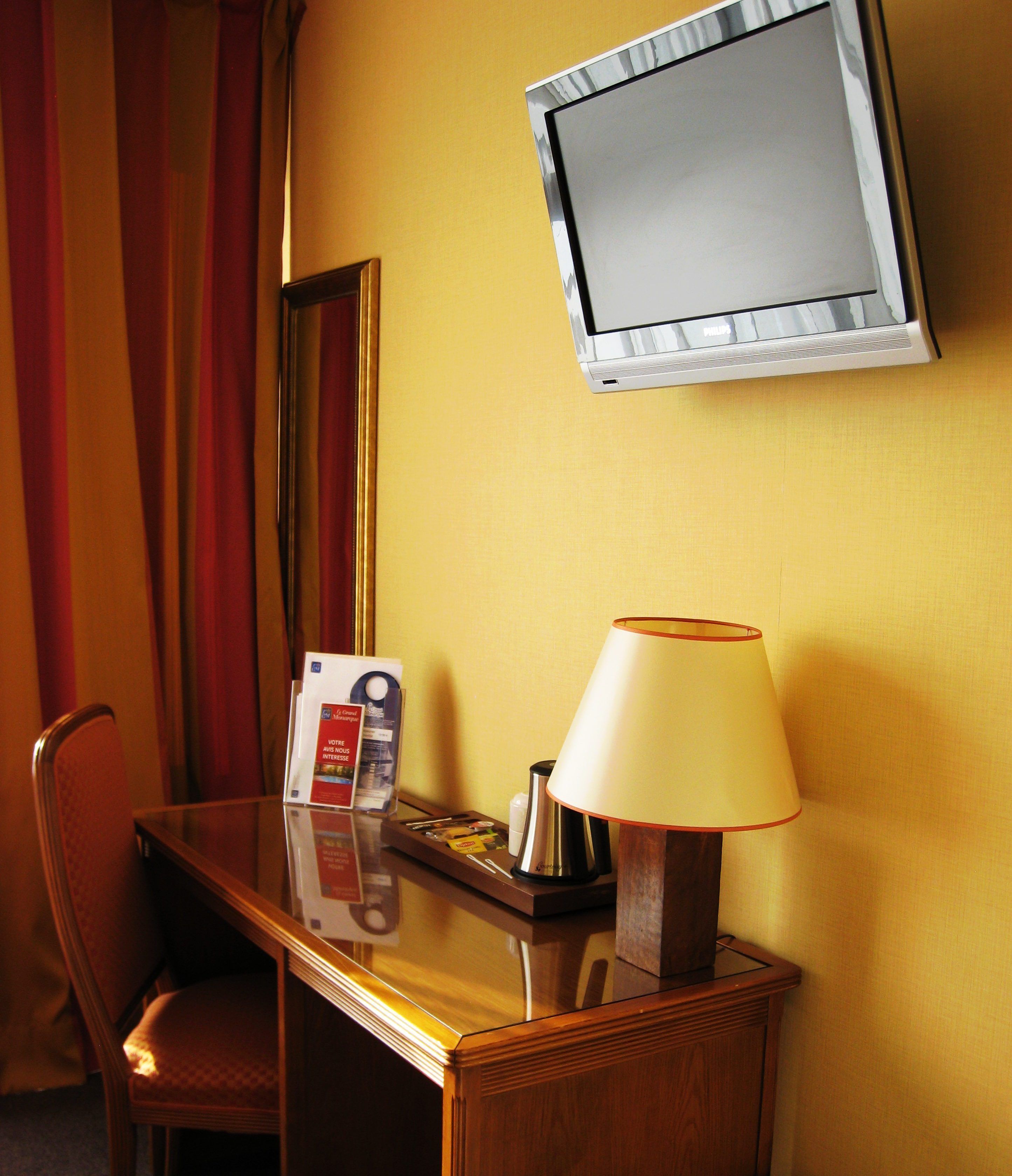 A hotel room with a wooden desk, a lamp, a television mounted on the wall, and a chair.
