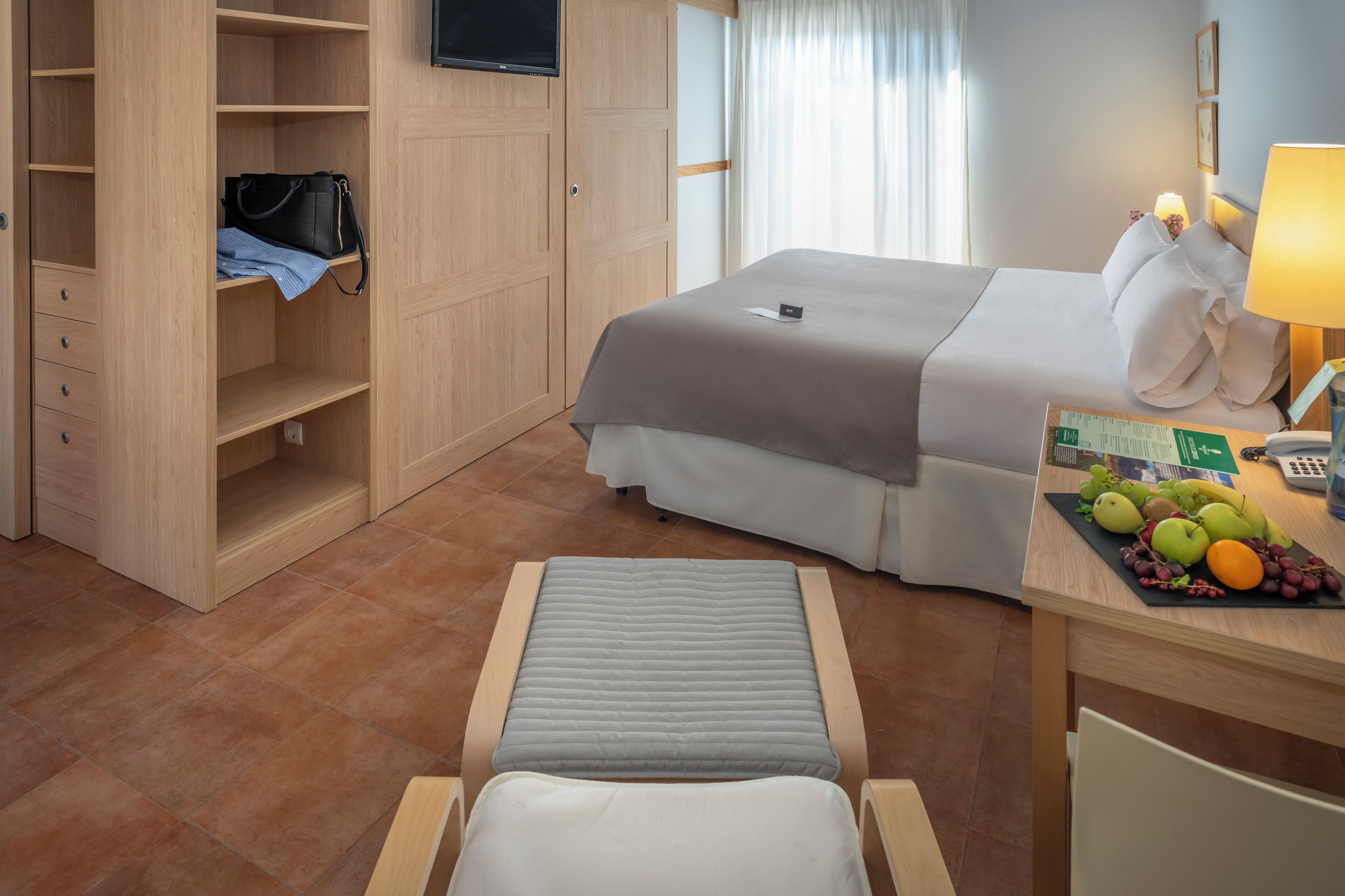 Habitación de hotel con cama, escritorio con lámpara, una silla, una mesita con frutas, armario y televisor en la pared.