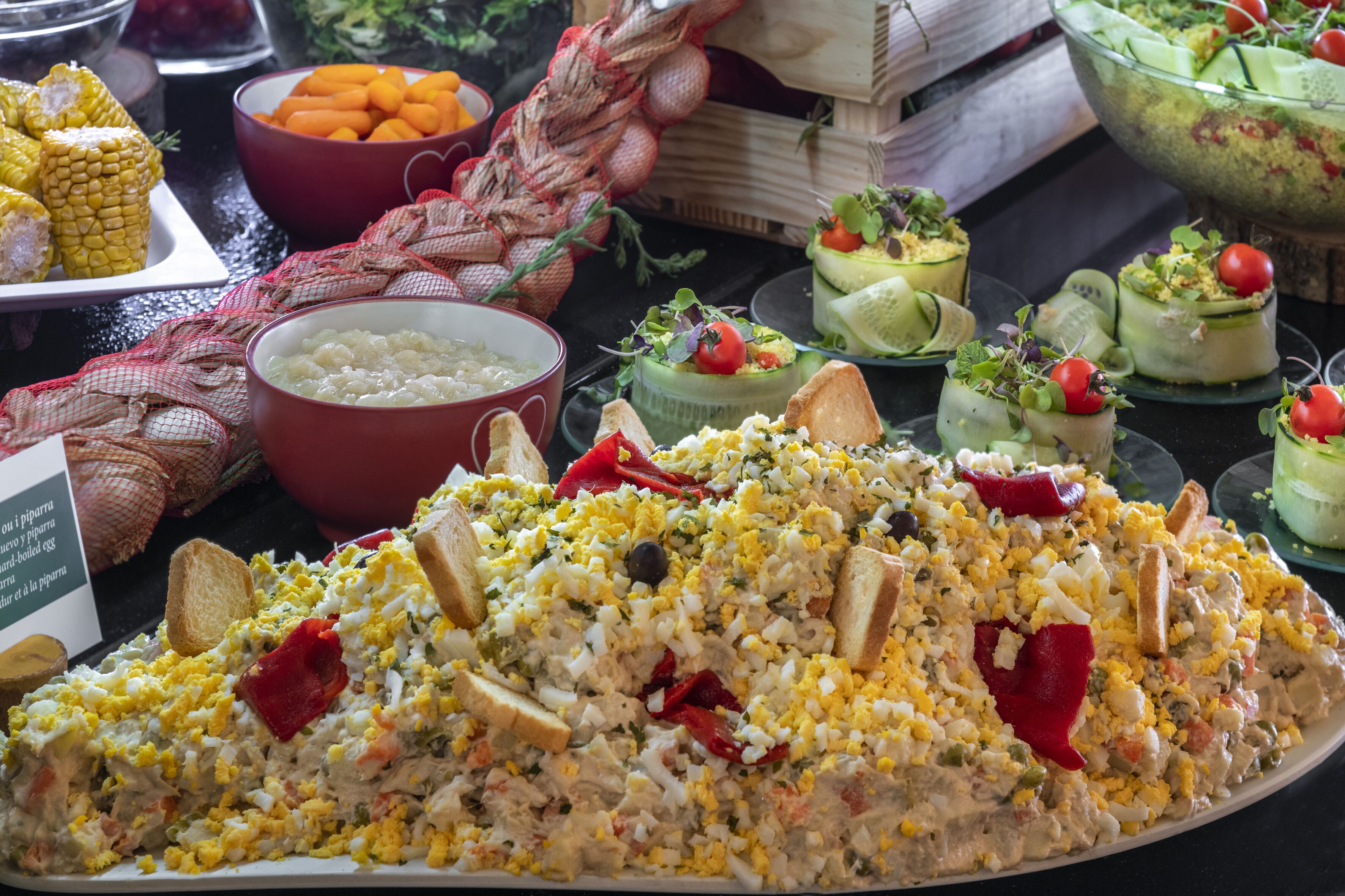 Una mesa de bufé con diversos platos, incluyendo una ensalada grande, mazorcas de maíz y otras comidas preparadas.