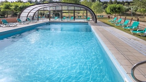 Una piscina con agua azul cristalina, rodeada de sillas de descanso verdes y un área de estar cubierta.