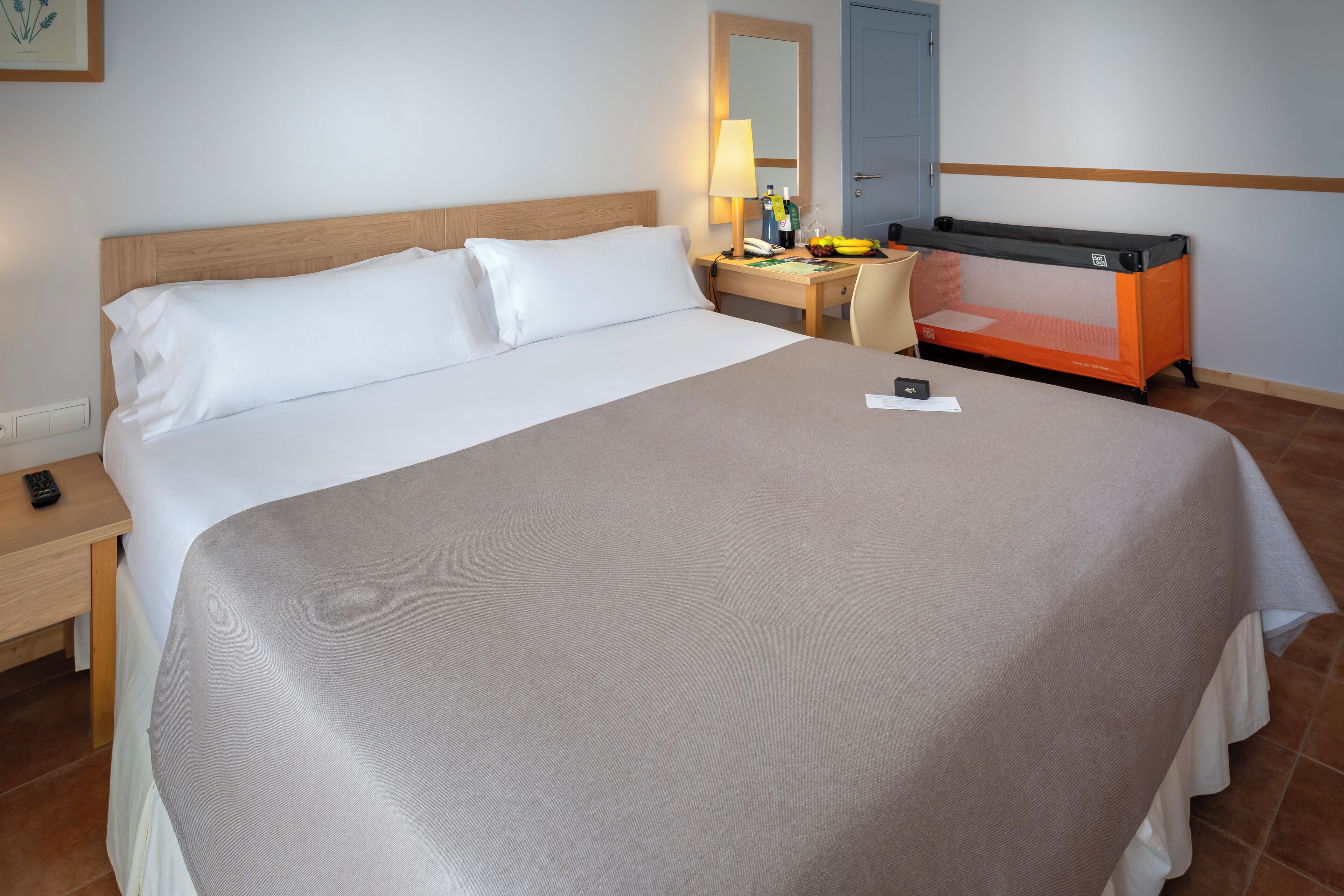 Habitación de hotel con una cama grande, un escritorio con lámpara, una silla y una pequeña mesa con una cesta de frutas.