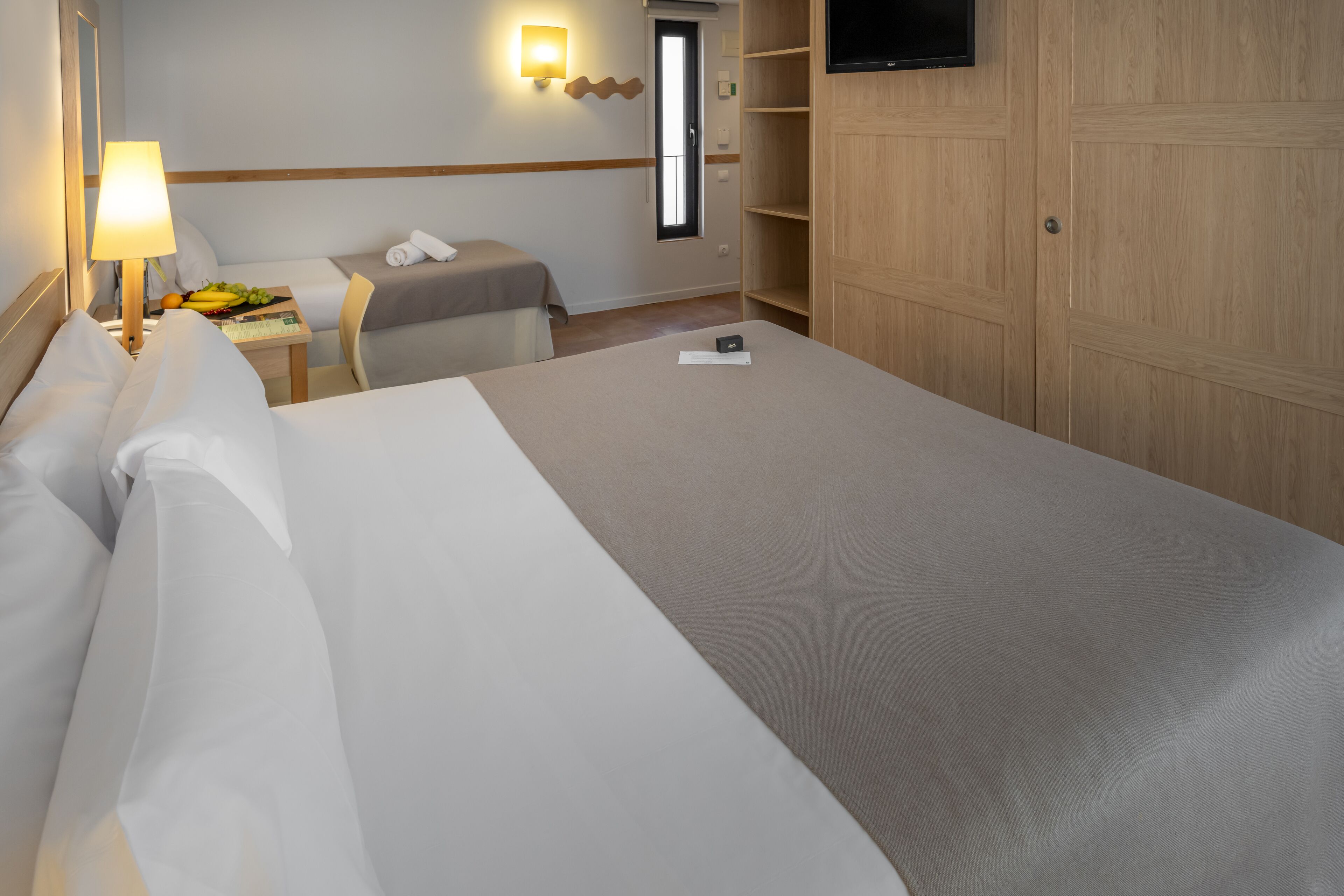 A hotel room with a large bed, a bedside table with a lamp, a small table with fruits, a TV on the wall, and a wardrobe.
