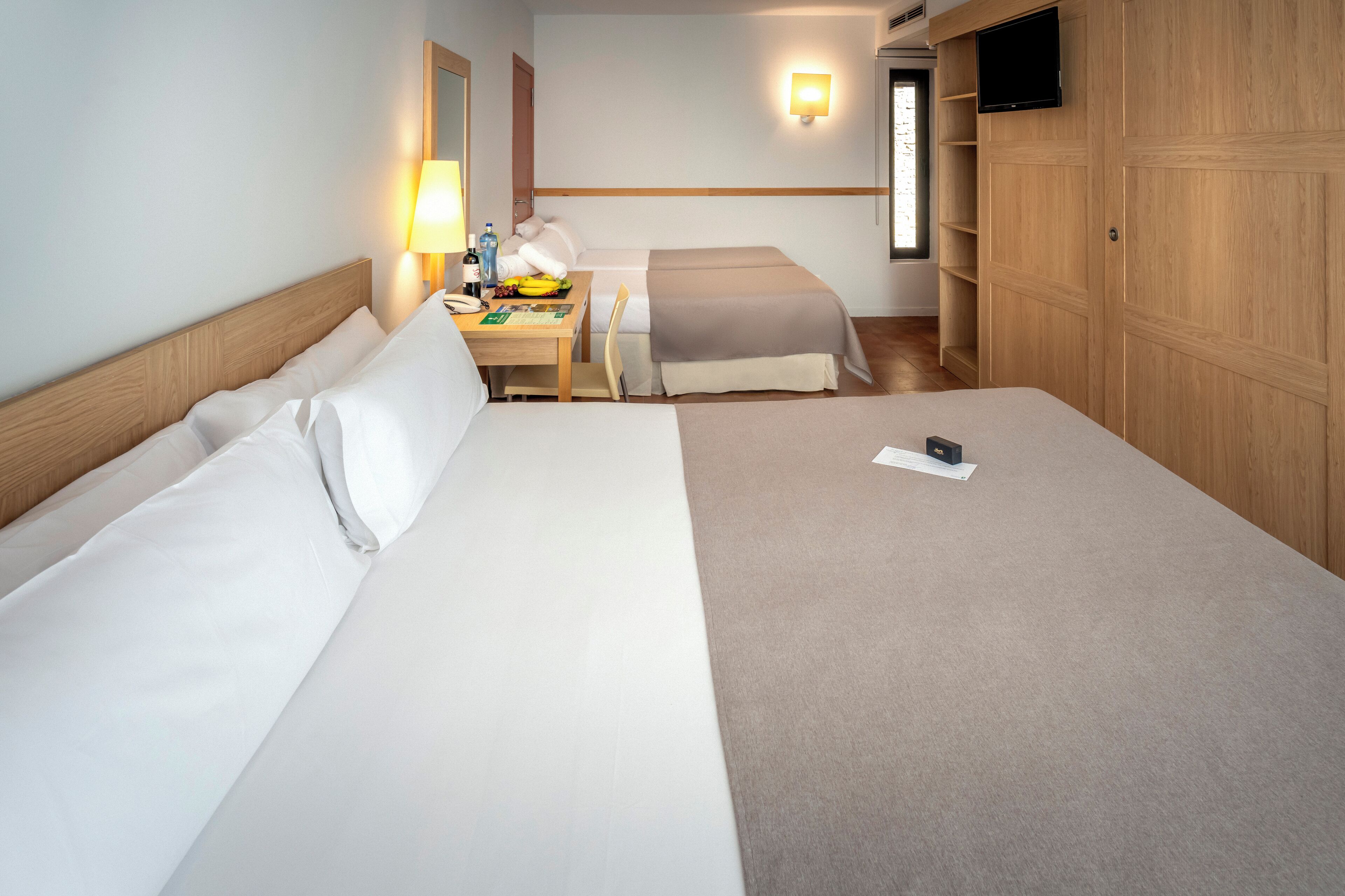 Habitación de hotel con dos camas, un escritorio con lámpara, televisor y armario.