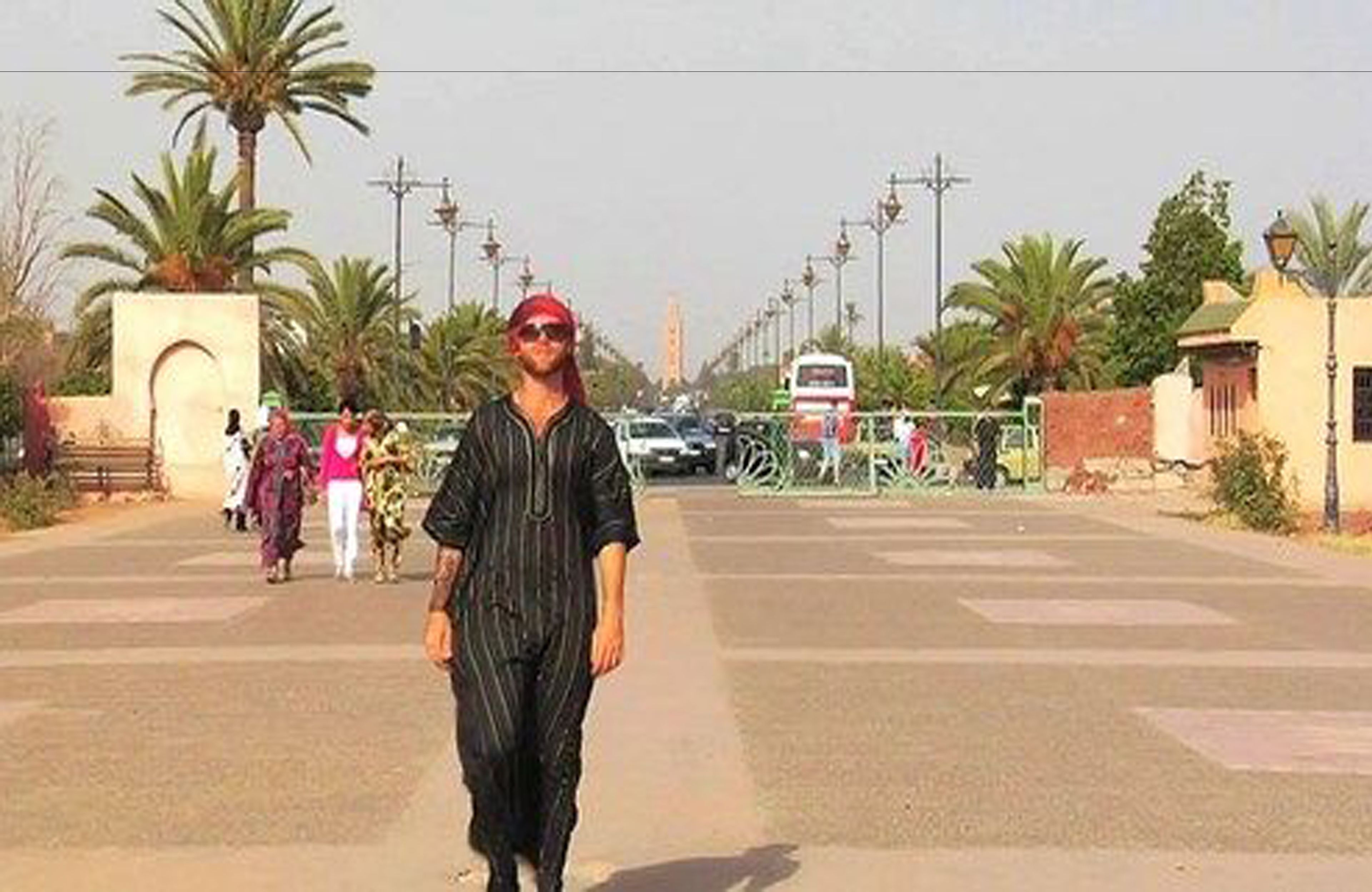 A man walking on a road with palm trees and a building in the background.