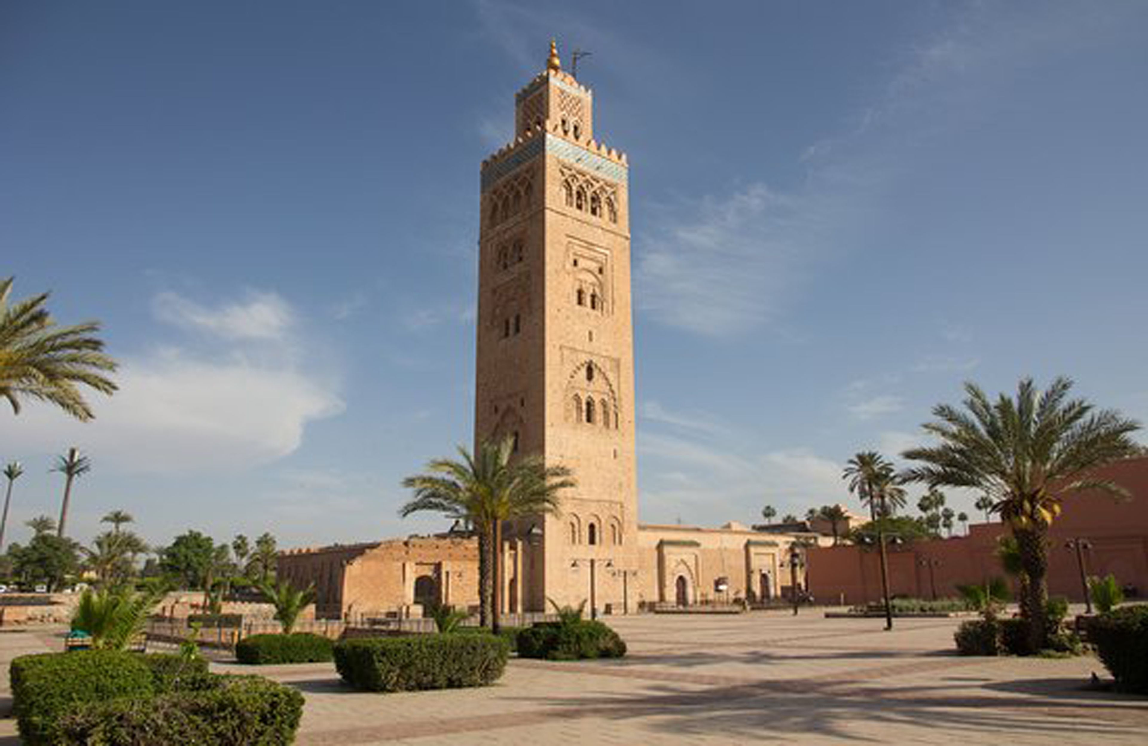 Ein hoher, beige farbener Turm mit aufwendigen Verzierungen, umgeben von einem geräumigen Innenhof mit Palmen und blauem Himmel.