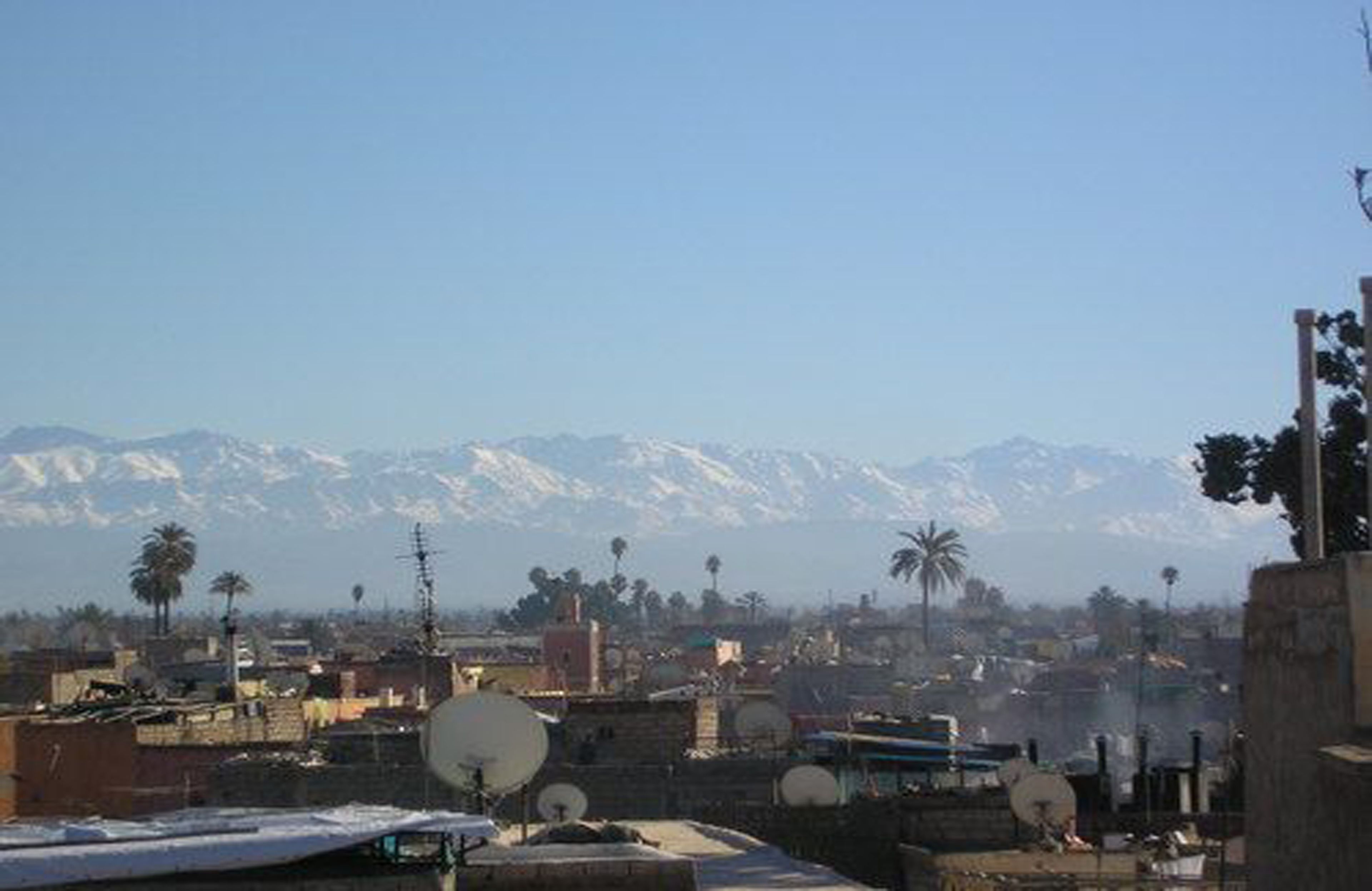 A cityscape with snow-capped mountains in the background, satellite dishes on rooftops, and palm trees.