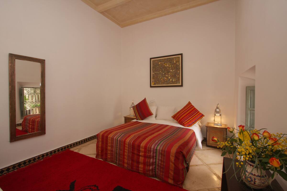 Quarto com cama grande, mesa de cabeceira, espelho, obra de arte emoldurada e um vaso com flores.