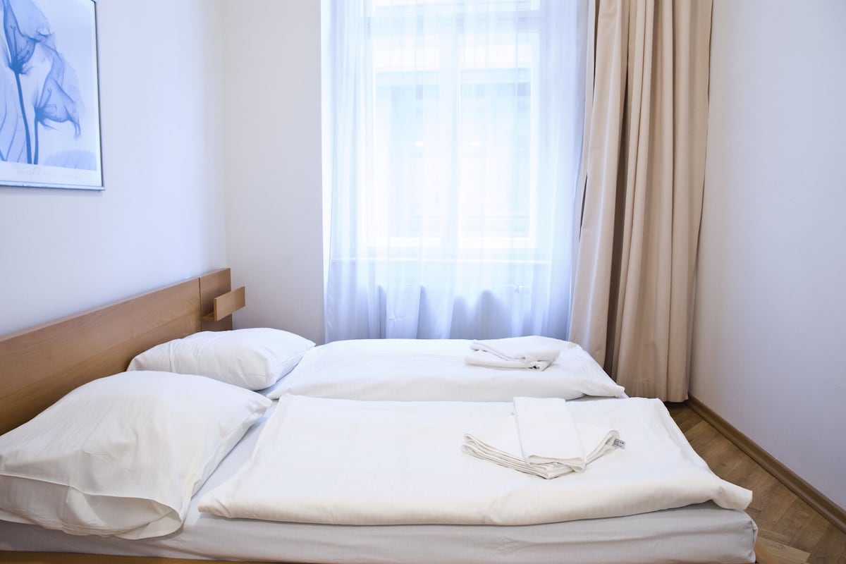 Ein ordentlich bezogenes Doppelbett mit weißen Leinen und einem gefalteten Handtuch, ein Holzkopfstück und ein Fenster mit transparenten Vorhängen.