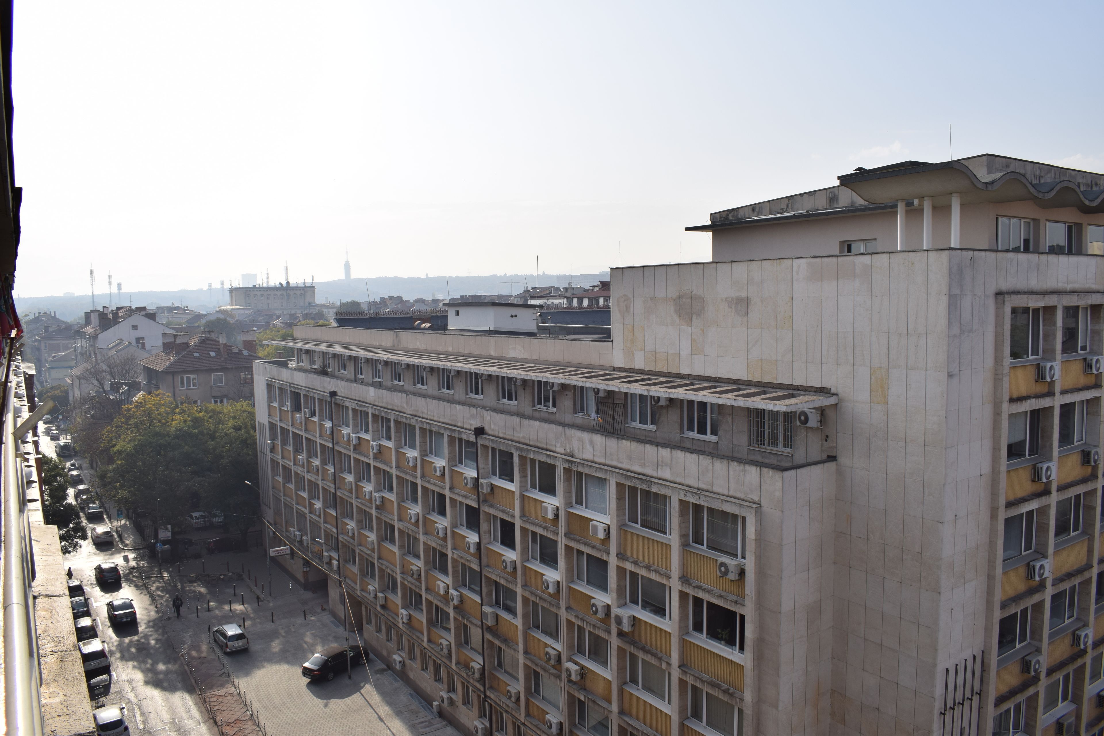 Un edificio de varios pisos con una cuadrícula de ventanas y balcones, un estacionamiento con varios automóviles y una vista despejada del paisaje urbano circundante.