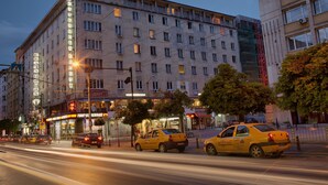 Et hotel med flere etager, et tydeligt skilt, forskellige butikker i stueplan og gule taxaer på gaden.