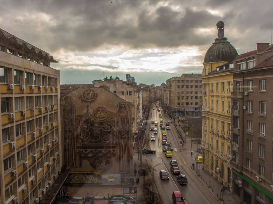 Una calle urbana flanqueada por edificios, una vía concurrida por automóviles y una gran estructura en forma de cúpula al fondo.