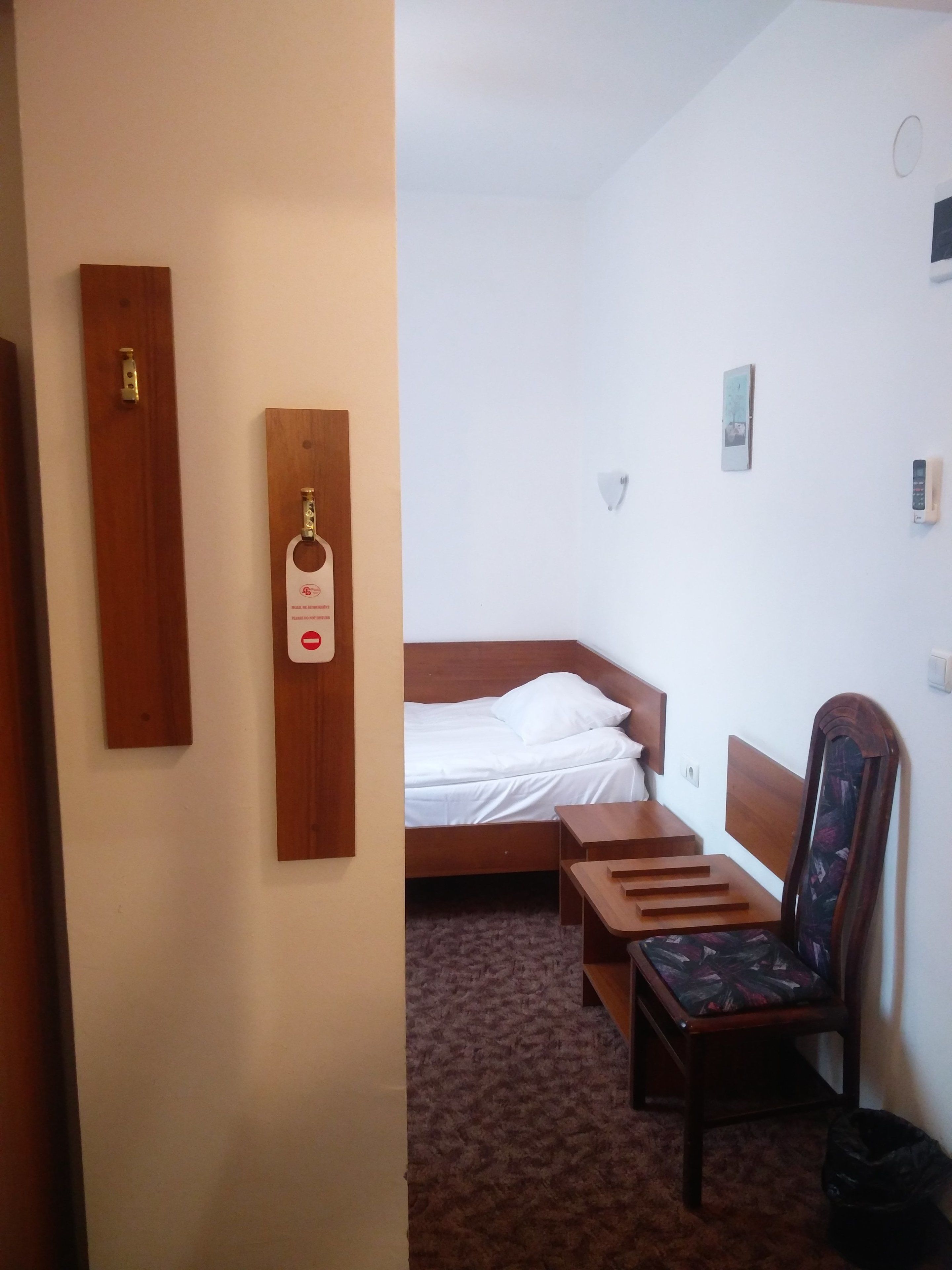 Una habitación de hotel con una cama, una silla de madera, una mesita pequeña y un gancho en la pared.