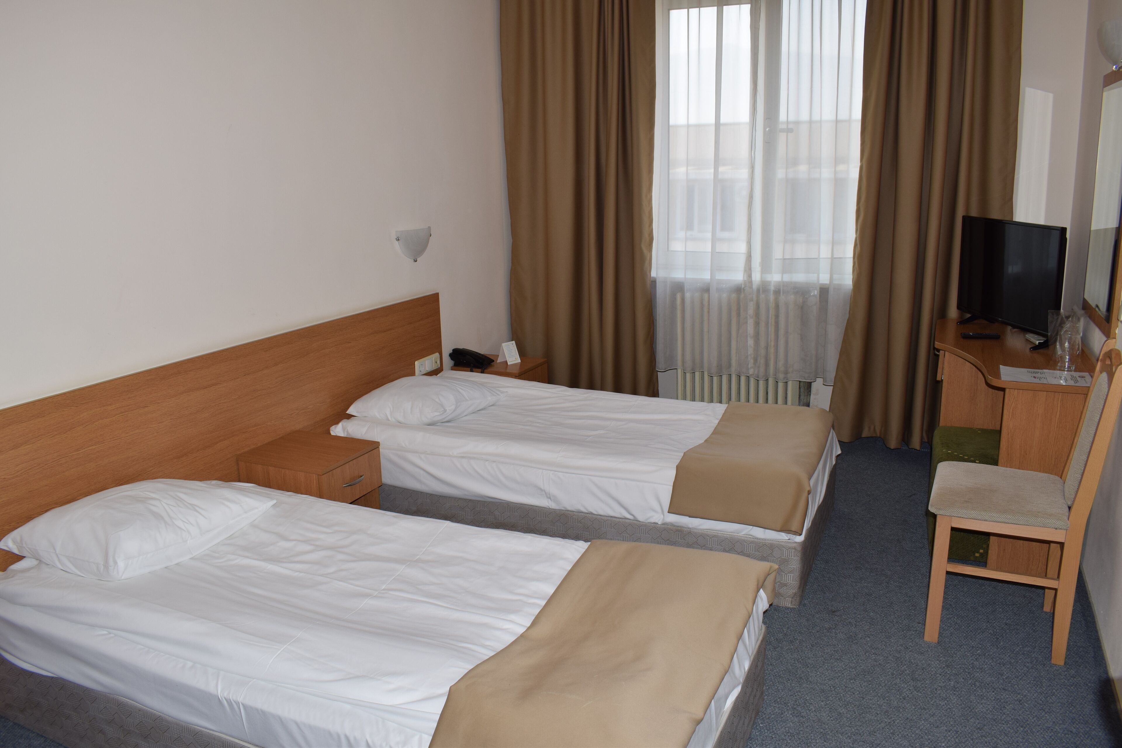 Et hotelværelse med to senge, et skrivebord med stol, et fjernsyn og et vindue med gardiner.