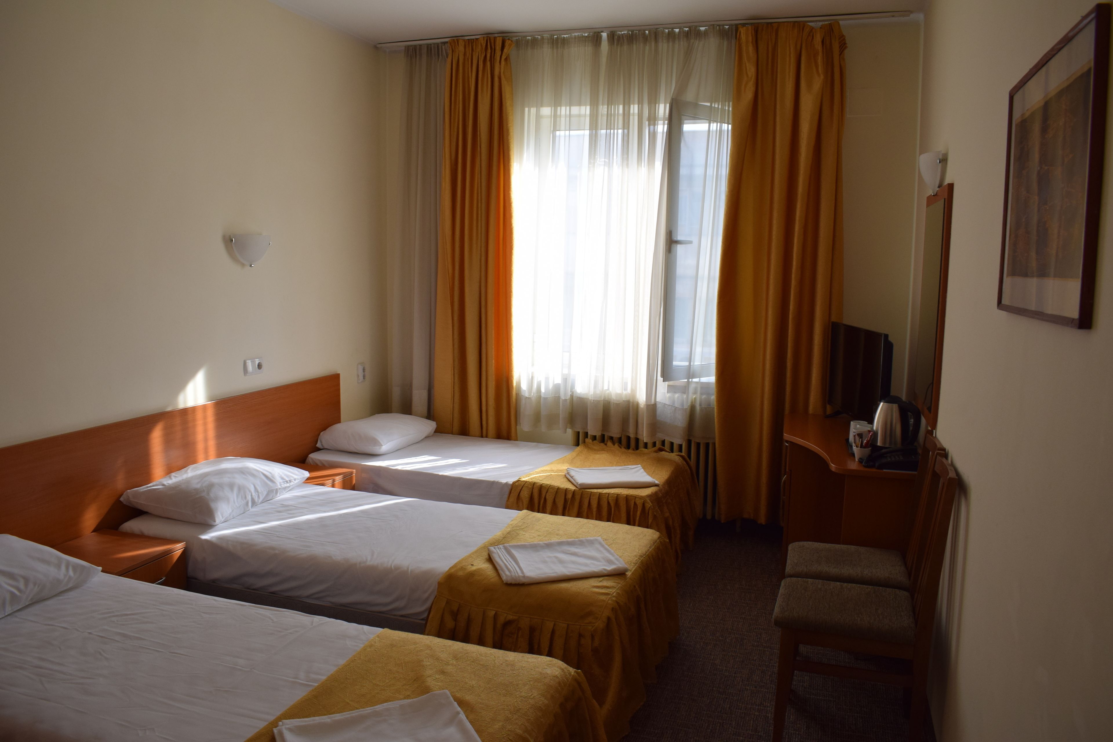 Ein Hotelzimmer mit drei Einzelbetten, einem Schreibtisch mit Stuhl, einem Fenster mit Vorhängen und einem gerahmten Bild an der Wand.