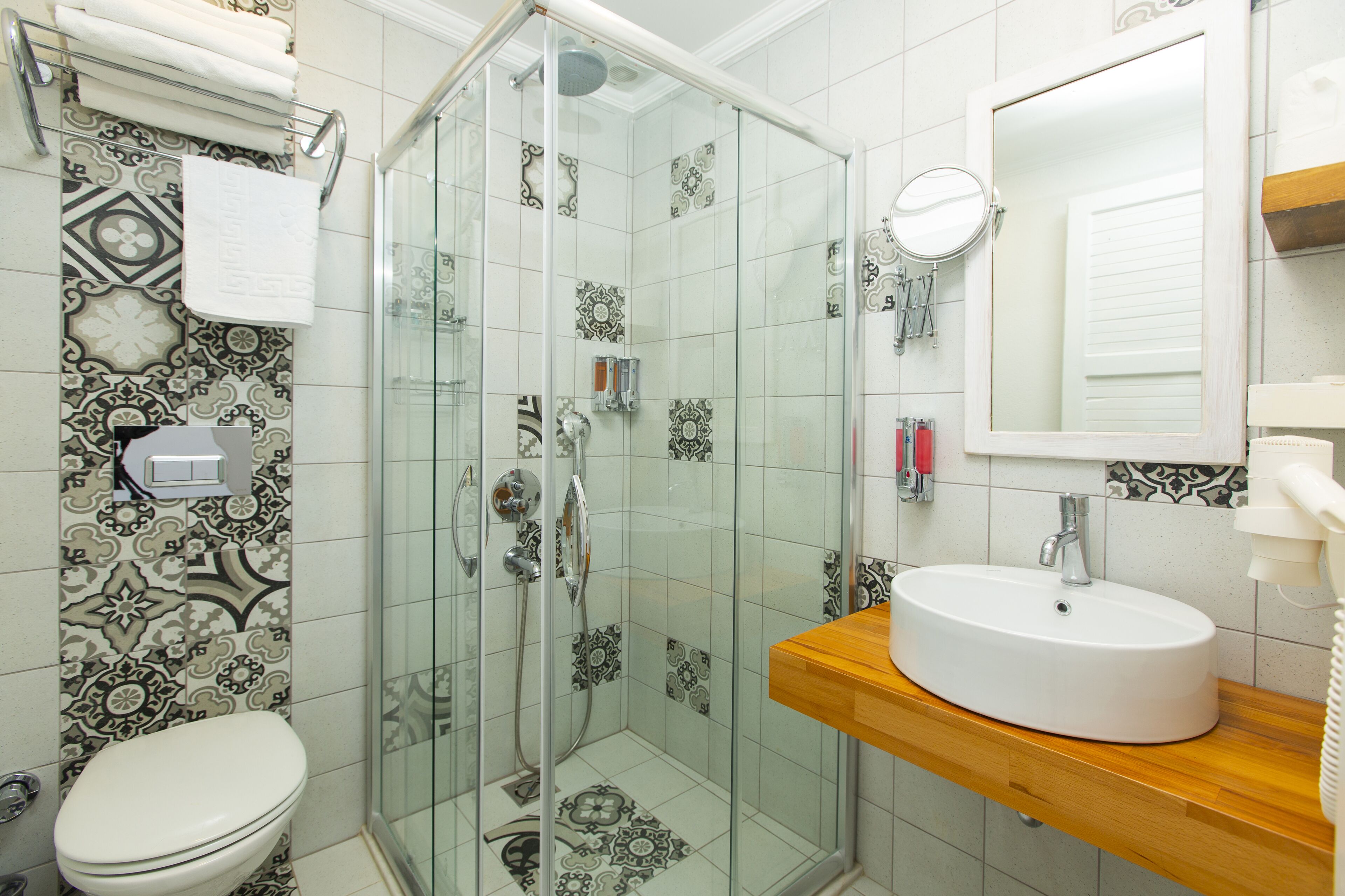 Un baño con ducha envuelta en vidrio, un inodoro blanco, un tocador de madera con lavabo blanco y azulejos estampados en las paredes.