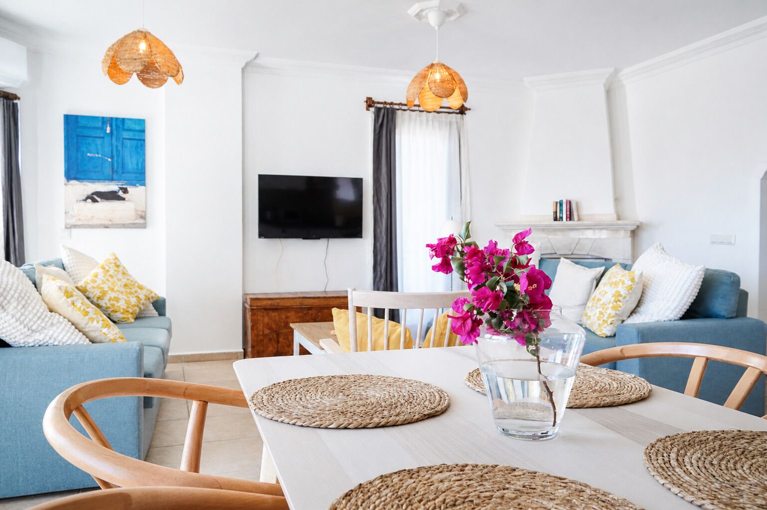 A living room with a white table, blue sofa, and wooden chairs. There is a TV mounted on the wall, a blue door, and a window with curtains.