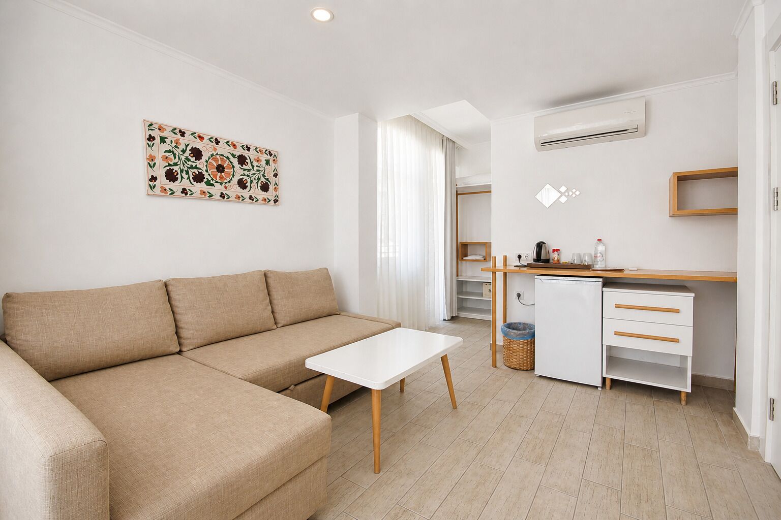 Una sala moderna con un sofá beige, una mesa blanca, una kitchenette con refrigerador y un aire acondicionado instalado en la pared.