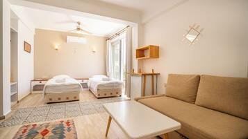 Una habitación moderna y bien iluminada con un sofá color beige, una mesa blanca y dos camas con ropa de cama blanca.