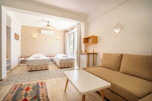 Una habitación moderna y bien iluminada con un sofá color beige, una mesa blanca y dos camas con ropa de cama blanca.