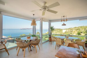 A spacious balcony with a table and chairs, large windows offering a view of the sea, and a ceiling fan.