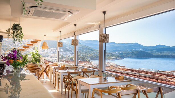 Un restaurante con terraza que da a un lago y montañas pintorescos, con mesas y sillas de madera y plantas colgantes.