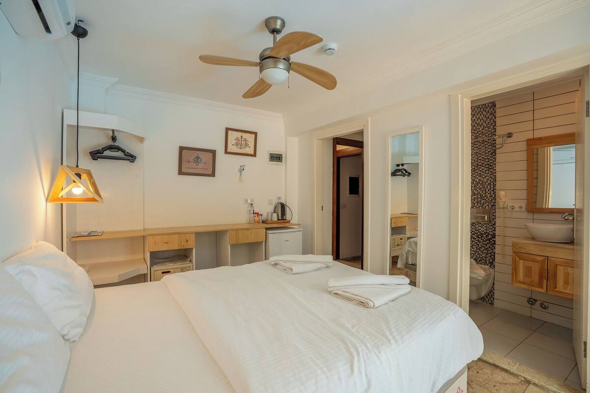 Habitación de hotel con cama, escritorio, ventilador de techo, lámpara de pared, refrigerador pequeño, baño con lavamanos y espejo, y una puerta que da a otra habitación.