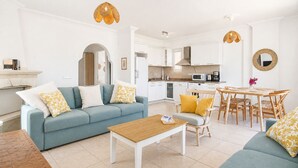 Una sala de estar moderna con sofá, mesa de centro, mesa de comedor y sillas. Hay una zona de cocina con armarios y electrodomésticos blancos.