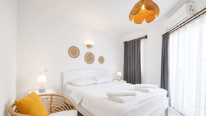 Habitación de hotel con una cama blanca, dos lámparas de noche, una silla de mimbre con un cojín amarillo y un gran ventanal con cortinas.