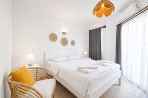 Habitación de hotel con una cama blanca, dos lámparas de noche, una silla de mimbre con un cojín amarillo y un gran ventanal con cortinas.