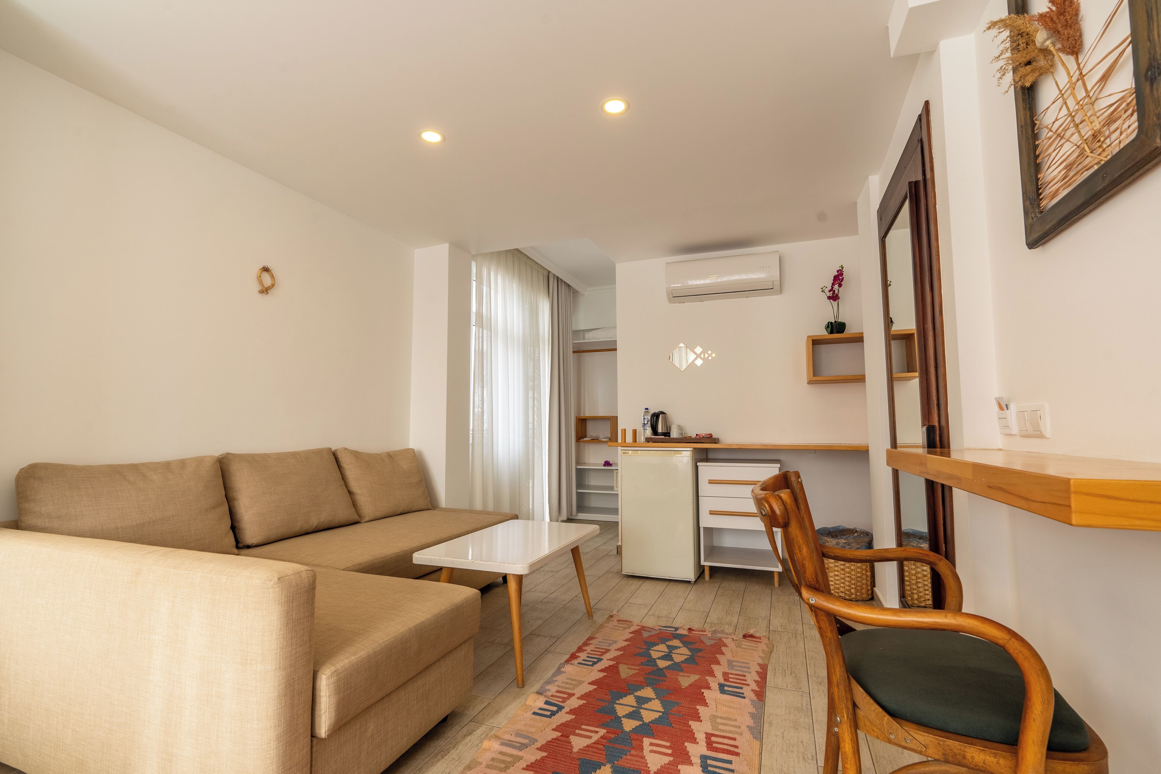 A modern living room with a sofa, a small table, and a wooden chair. There is a kitchen area with a refrigerator and a sink.