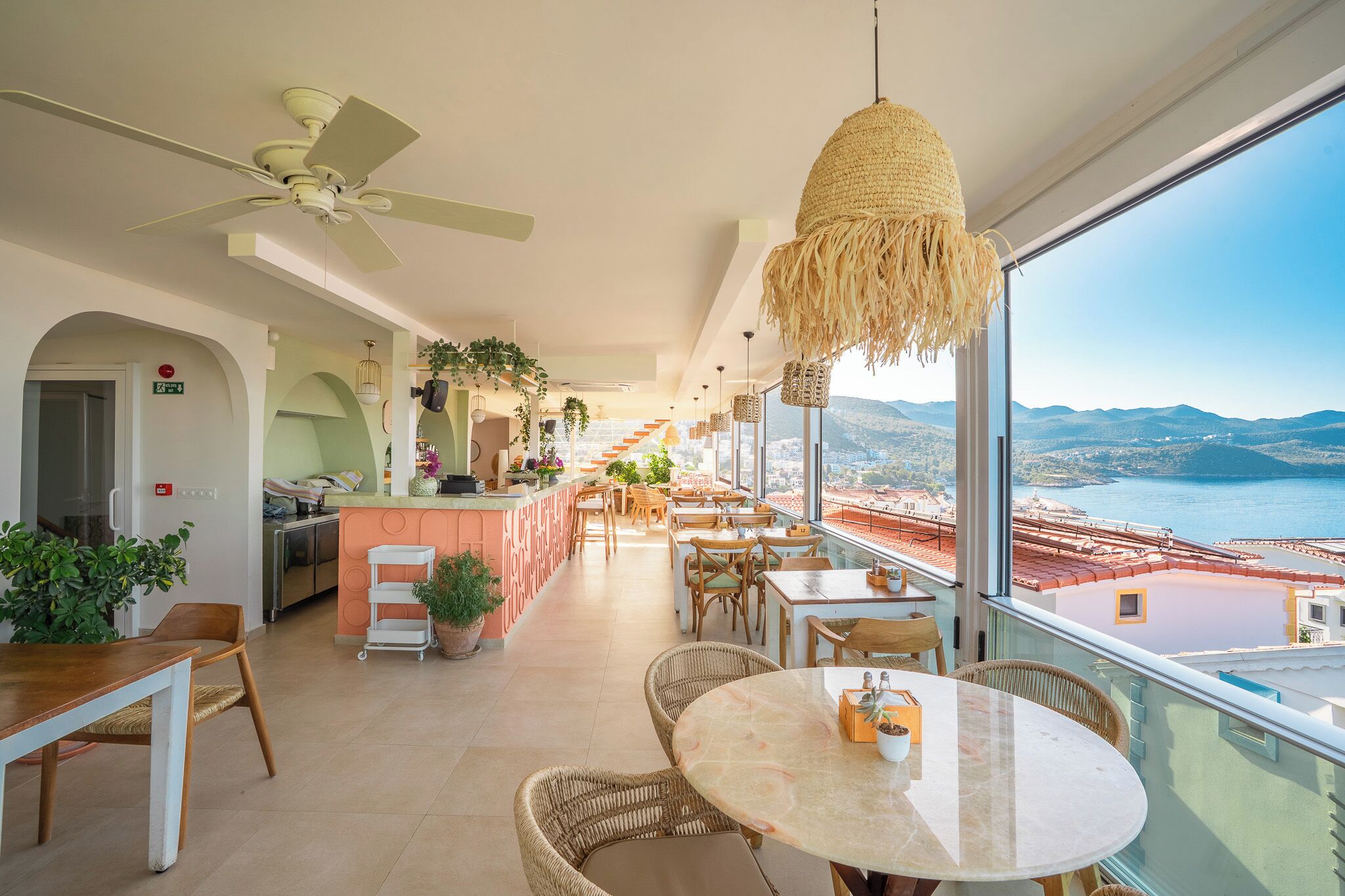 A spacious balcony with a view of the sea and mountains, furnished with tables and chairs, and a ceiling fan.