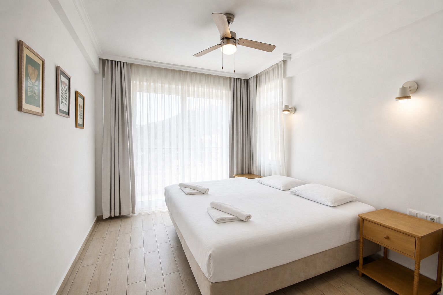 Une chambre d’hôtel avec un lit, une table de chevet, un ventilateur de plafond et une fenêtre avec des rideaux.