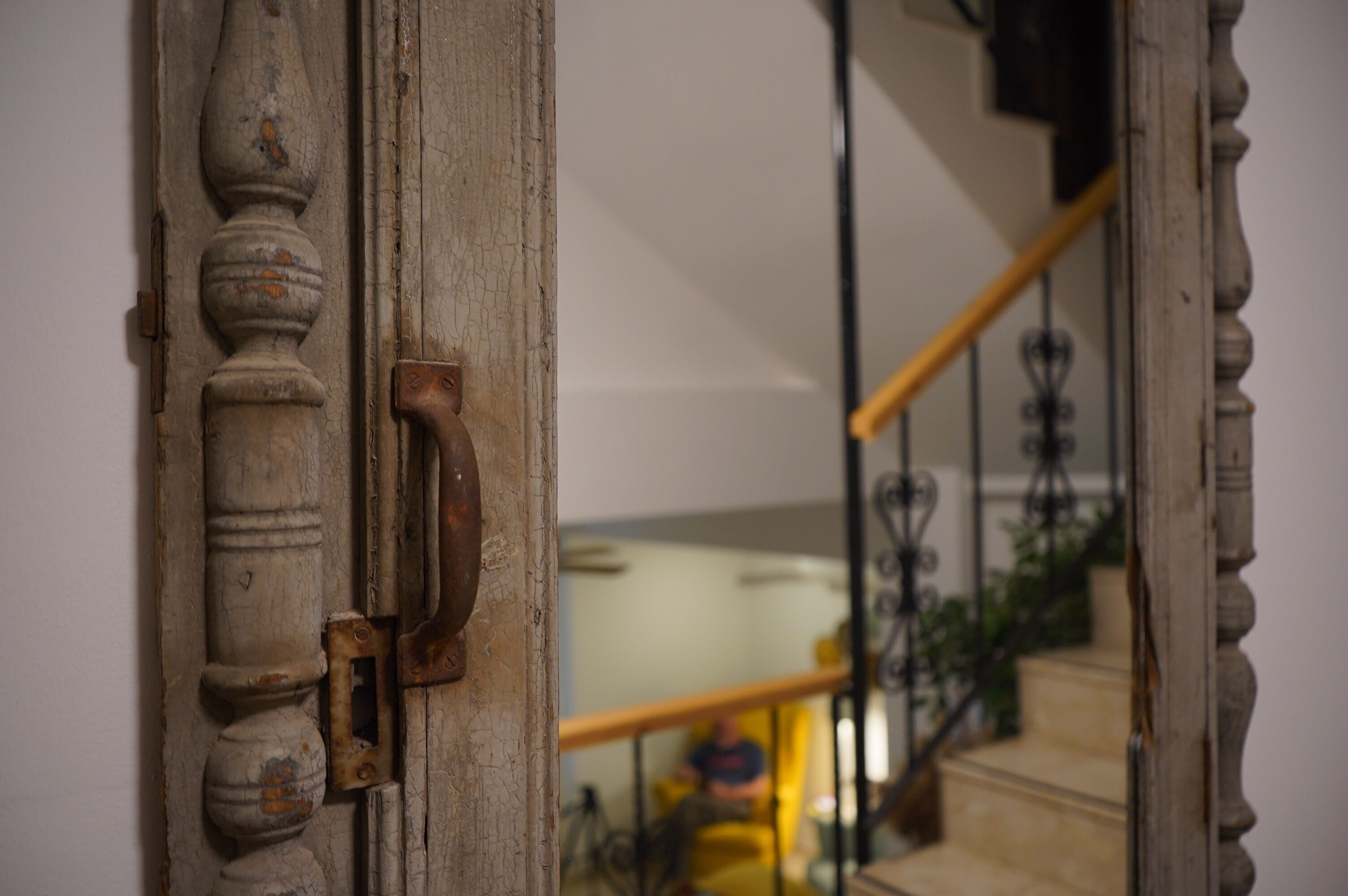 A close-up of a weathered wooden door with a brass handle, leading to a staircase with a metal railing and a yellow chair in the background.