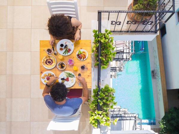 A dining area with a pool view, featuring a wooden table with plates of food, white chairs, and potted plants.