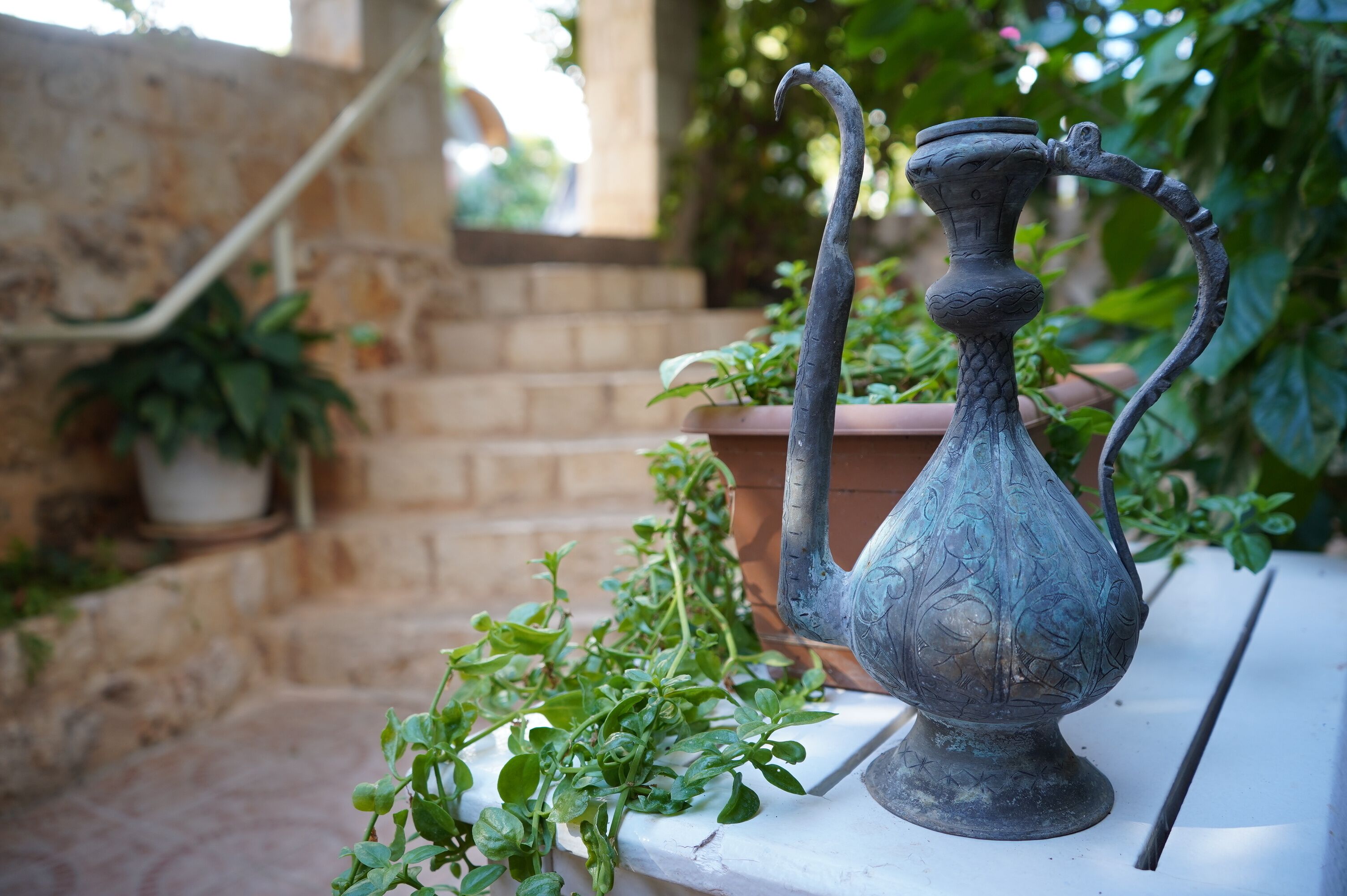 A metal vase with a curved handle on a white table in an outdoor setting with potted plants and a stone wall.
