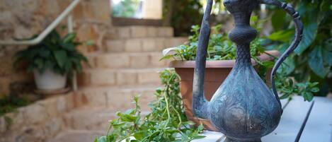 A metal vase with a curved handle on a white table in an outdoor setting with potted plants and a stone wall.