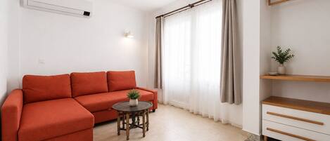 Una sala de estar moderna con un sofá, una mesita redonda con una planta, un aire acondicionado de pared, una ventana con cortinas traslúcidas y un estante de madera con una planta.