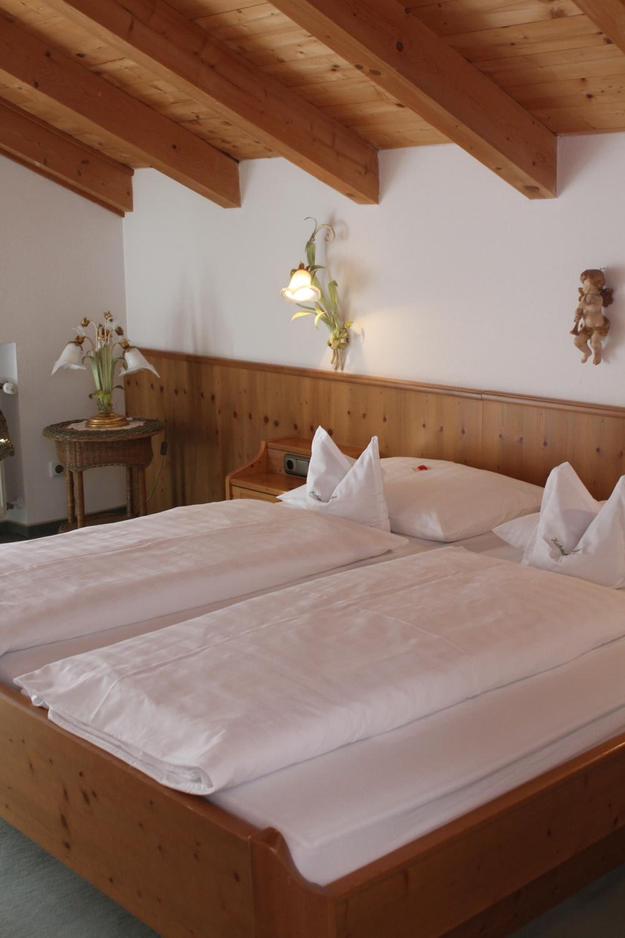 A double bed with white linens, a wooden headboard, and a small bedside table with a lamp and a vase of flowers.