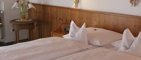 A double bed with white linens, a wooden headboard, and a small bedside table with a lamp and a vase of flowers.