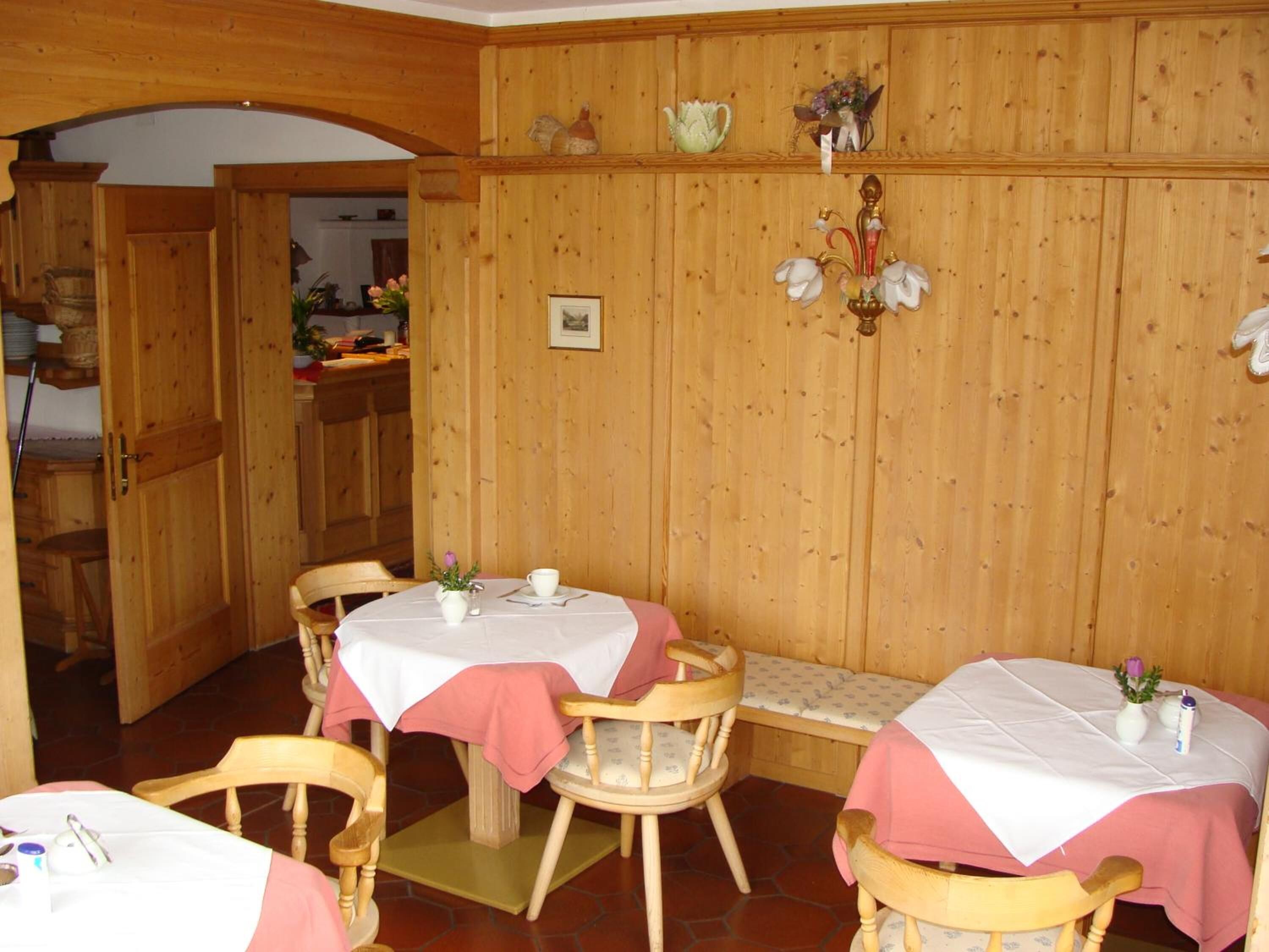 Ein gemütlicher Essbereich mit Holzwänden und einer Bank. Es sind zwei Tische mit weißen Tischdecken und rosa Tischläufern aufgestellt. Eine Deckenleuchte hängt von der Decke, und ein gerahmtes Bild ist an der Wand zu sehen.