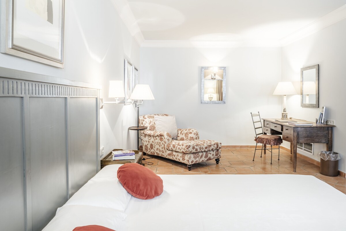 A hotel room with a bed, a patterned armchair, a desk with a chair, and a small table with books.