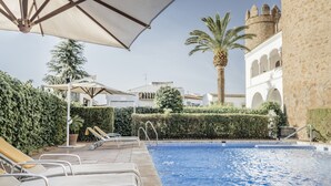 Zona de piscina con sillones de descanso, sombrillas y una palmera.