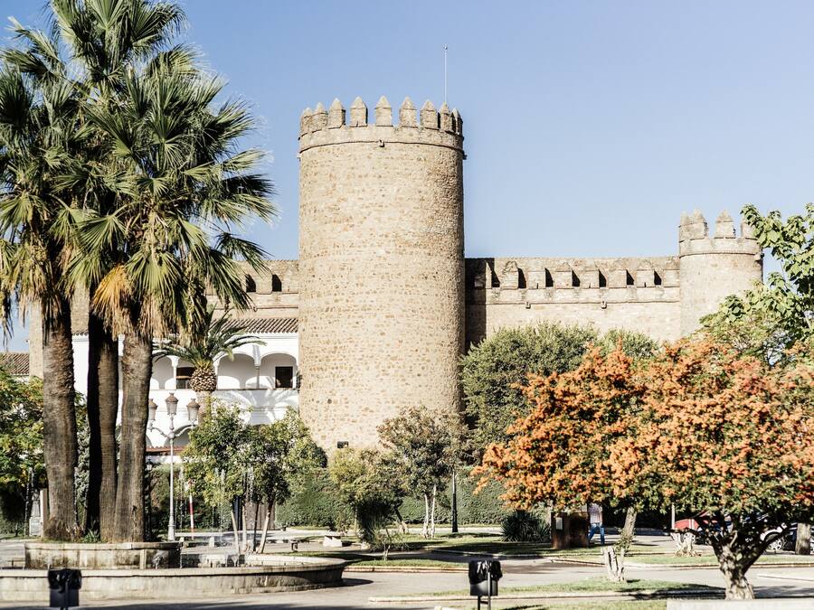 Una torre de piedra histórica con almenas, rodeada de palmeras y un parque con una fuente.