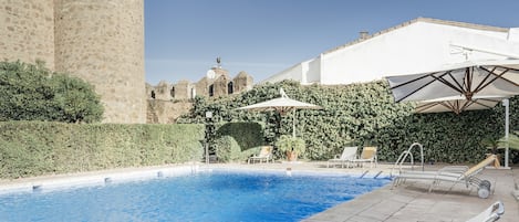 Una piscina con sillones de descanso y sombrillas, rodeada de follaje y antiguas paredes de piedra.