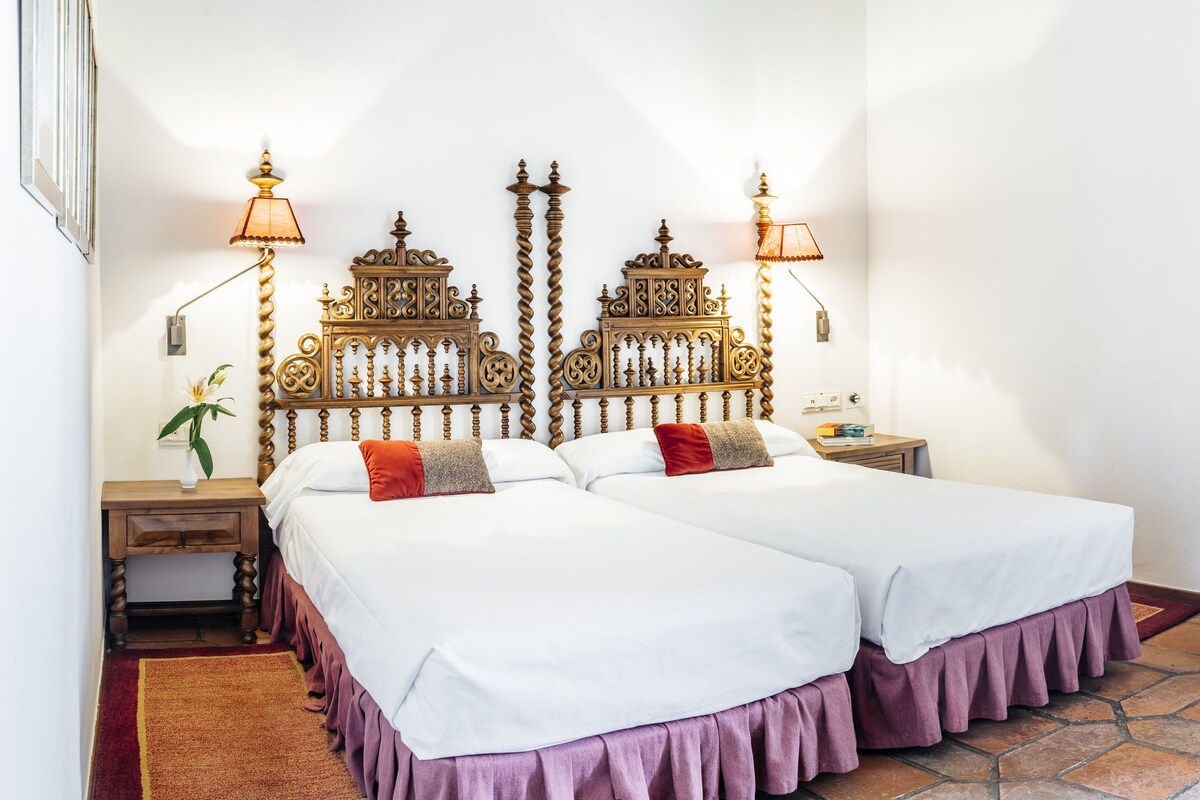 A double bed with a wooden headboard, two bedside tables, a nightstand, a lamp, and a curtain.