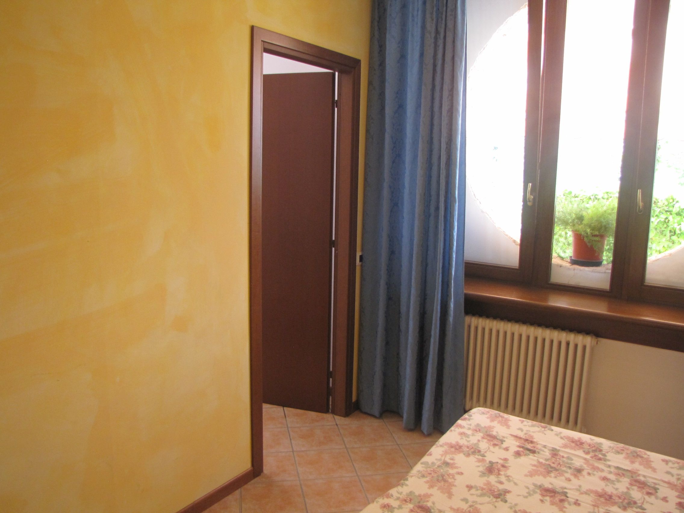 Una habitación con cama, ventana con cortinas, radiador y puerta que da a otra estancia.